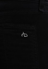 Jeans en denim noir avec une poche arrière arborant un logo brodé blanc. Le tissu a une texture lisse et un design à coupe classique.