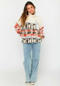 Veste en polaire crème avec des accents en rouge et noir. Présente un col montant, une fermeture éclair et une poche poitrine avec une bordure bleue.