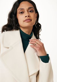 Krageförsedd, krämfärgad ullrock med dubbelknäppning, kombinerad med en grön polotröja. Synliga guldörhängen med hoop-design och en texturerad ring.