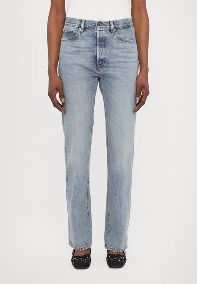 Hellblaue Jeans aus Denim mit geradem Schnitt, fünf Taschen, subtilen Verwaschungen und Nähdetails; getragen mit schwarzen flachen Schuhen.