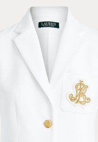 Sacou alb cu monogram "LR" brodat cu fir auriu pe buzunar, nasture auriu și etichetă "Lauren Ralph Lauren" în interiorul gulerului.