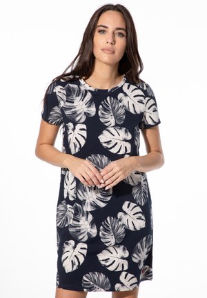 Frau in einem marineblauen kurzärmligen Kleid mit großem weißem tropischem Blattmuster, die mit locker verschränkten Händen vor sich steht.