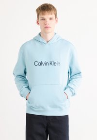 Svetlo modrá mikina s kapucňou vyrobená z bavlnenej zmesi. Obsahuje predné vrecko, elastické manžety a výrazné logo "Calvin Klein" v tmavomodrej farbe.