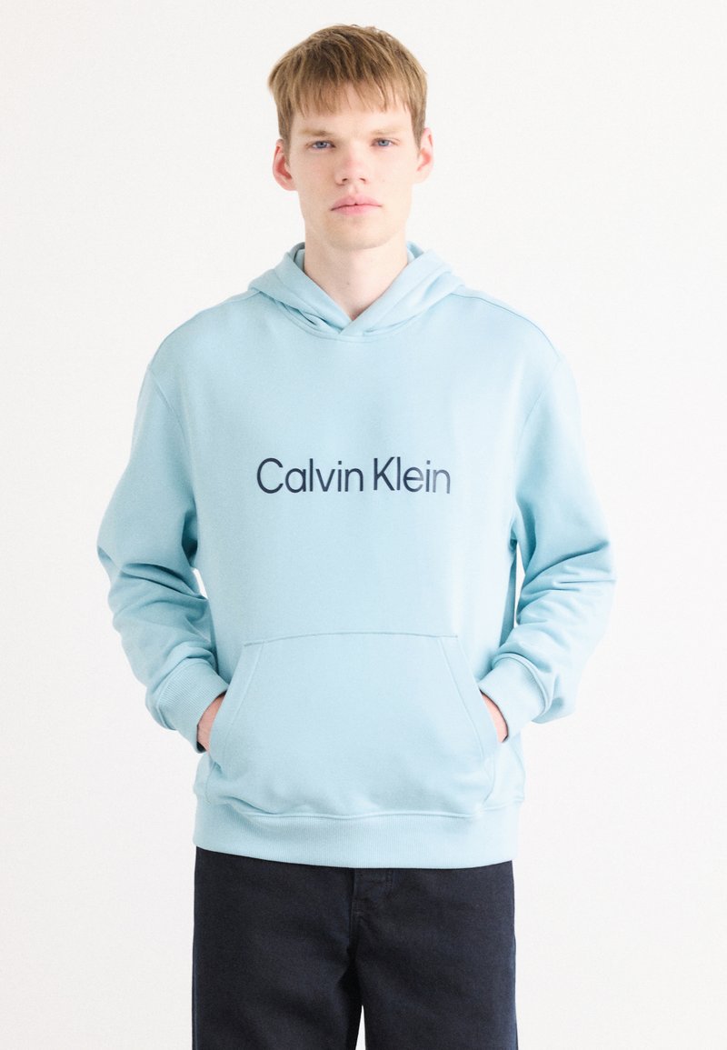 Svetlo modrá mikina s kapucňou vyrobená z bavlnenej zmesi. Obsahuje predné vrecko, elastické manžety a výrazné logo "Calvin Klein" v tmavomodrej farbe.