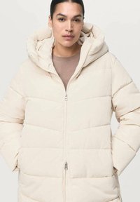 Lichtbeige pufferjack met een capuchon, voorzien van een rits aan de voorkant en horizontale stiksels. De stof lijkt glad en zacht van textuur.