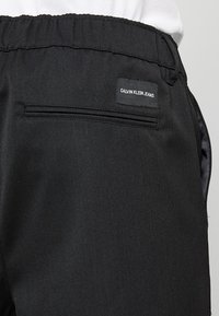 Černé kalhoty z látky se hladkým povrchem, s elastickým pasem a zadní kapsou se šitým štítkem s nápisem "CALVIN KLEIN JEANS."