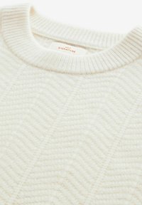 Pull en tricot couleur crème avec un col rond côtelé, arborant un motif chevron texturé et une étiquette portant la mention "NEXT SIGNATURE."