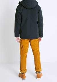 Chaqueta negra aislante con capucha, combinada con pantalones cargo de pana mostaza que tienen bolsillos laterales, y botas marrones con suelas blancas.