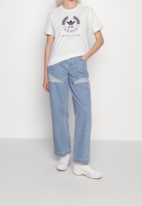 Witte katoenen t-shirt met marineblauw grafisch print en tekst. Lichtblauwe spijkerbroek met contrasterende stofpatches. Witte sneakers maken de outfit compleet.