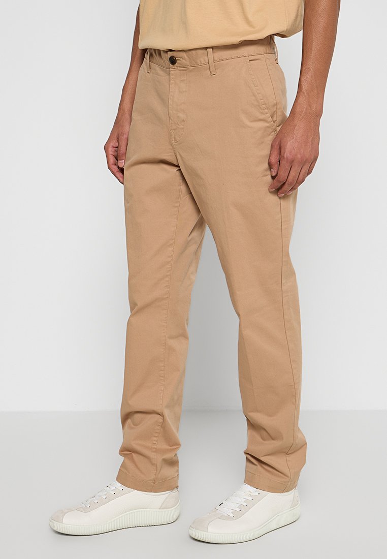 Beige Baumwoll-Chinohosen mit geradem Bein, ausgestattet mit Seitentaschen und flachem Bund, kombiniert mit weißen Sneakern.