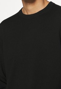 Homme portant un pull noir uni à col rond, montrant la partie supérieure de la poitrine et le cou.