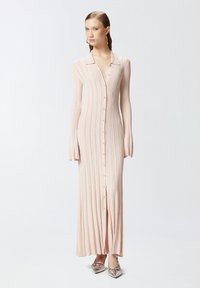 Duga, rebrasta haljina svijetlo ružičaste boje s ovratnikom, prednjim dugmadi i širenjem na rukavima; uparena s metalik cipelama s oštrim vrhom.