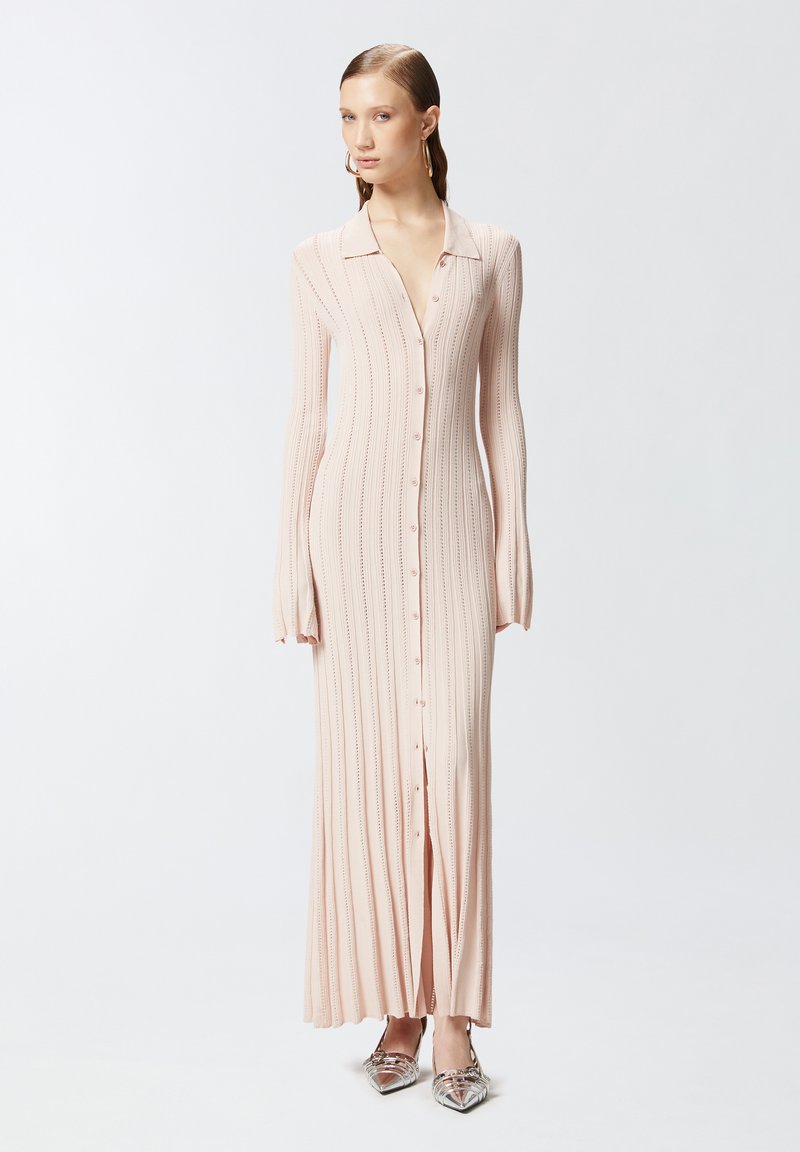 Duga, rebrasta haljina svijetlo ružičaste boje s ovratnikom, prednjim dugmadi i širenjem na rukavima; uparena s metalik cipelama s oštrim vrhom.