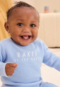 Svetle modrý pletený overal s bílým logem, dlouhými rukávy a knoflíkovými detaily na rameni. Měkká textura, kulatý výstřih a pohodlný střih.