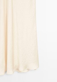 Robe de couleur crème en tissu texturé, avec un design fluide qui descend jusqu'au sol. Aucuns motifs visibles ni détails matériels.