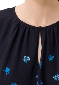 Blouse navy avec décolleté froncé, motifs floraux bleus brodés, ouverture en clef à l'avant et un petit détail bouton. Tissu léger.