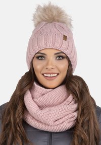 Roze gebreide beanie met een lichte bont pompon bovenop, gekenmerkt door een dikke ribstructuur. Gecombineerd met een bijpassende gebreide infinity sjaal.