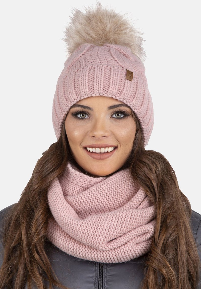 Roze gebreide beanie met een lichte bont pompon bovenop, gekenmerkt door een dikke ribstructuur. Gecombineerd met een bijpassende gebreide infinity sjaal.