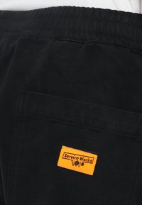 Zwarte broek van stof, met een elastische tailleband en een achterzak met een oranje label "Service Works". Gladde textuur.