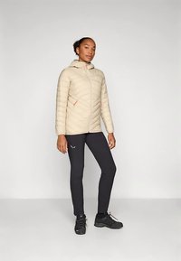 Šviesiai smėlio spalvos pūsta striukė su gobtuvu, diagoniškai siuvinėta, su oranžiniais akcentais. Derinama su juodomis sportinėmis tamprėmis ir batais.