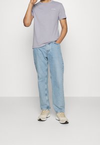 Camiseta de manga corta color lila, con logo en el pecho, combinada con jeans rectos color azul claro y zapatillas beige, de pie sobre un fondo neutro.