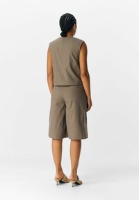 Bruine mouwloze top gecombineerd met knielange shorts, beide gemaakt van een gladde stof. Lichtbruine slippers met een lage hak maken de outfit compleet.