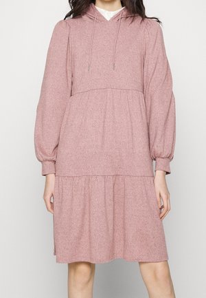 Kvinde iført en langærmet, lyserød, hætteklædt, lagdelt kjole med snoretræk og rynkede manchetter, stående mod en ensfarvet baggrund.