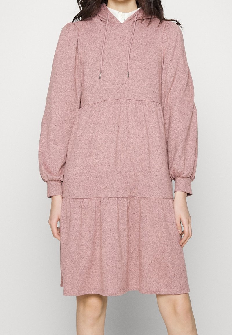 Femme portant une robe longue à manches longues, rose, à capuche, à volants avec des cordons et des poignets froncés, debout devant un fond uni.