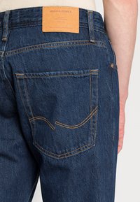 Jeans in denim blu scuro con un design distintivo della tasca posteriore, etichetta in pelle beige, cuciture arancioni e una silhouette aderente.