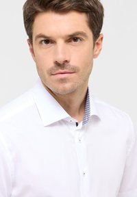 Camicia bianca con colletto strutturato e dettagli a motivo blu, chiusura frontale con un bottone e una texture liscia.