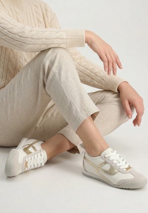 Osoba siedząca ze skrzyżowanymi nogami, ubrana w beżowe spodnie, kremowy sweter z warkoczowym splotem i białe trampki ze złotymi akcentami.