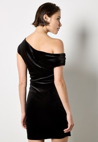 Abito in velluto nero con spalle scoperte, silhouette aderente, texture liscia e dettaglio di tessuto drappeggiato sulla spalla, design alla lunghezza del ginocchio.