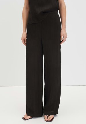 Vrouw die zwarte, hooggetailleerde wijde broek draagt, gecombineerd met een zwart mouwloos topje en zwarte teenslippers tegen een witte achtergrond.