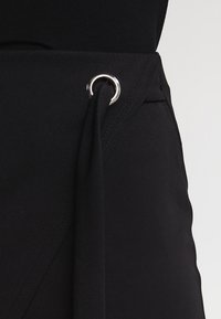 Černá sukně s hladkým povrchem, představující design s překrýváním a kovovým očkem pro nastavitelné zavázání.