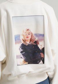 Femme blonde aux cheveux bouclés dans une veste sombre pose en extérieur, imprimée au dos d’un sweat-shirt blanc.