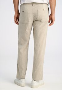 Lichtbeige gestreepte broek met een rechte snit, voorzien van twee achterzakken en gouden details. Draagt met witte sneakers.
