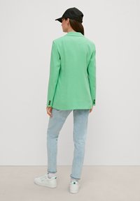 Grüne Blazer mit Reverskragen, Ein-Knopf-Manschetten und bequemer Passform, kombiniert mit hellblauen Jeans und weißen Turnschuhen.