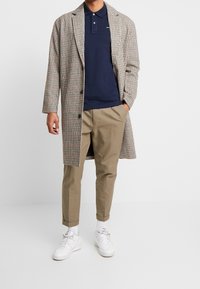 Man wearing navy polo, beige checked long coat, khaki pants, white socks, and white sneakers standing with one hand in pocket.