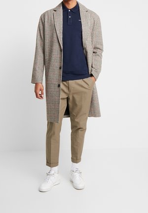 Mies pukeutuneena tummansiniseen poolopaitaan, beigeen ruudulliseen pitkässä takkiin, khakihousuihin, valkoisiin sukkiin ja valkoisiin tennareihin, seisomassa yhdellä kädellä taskussa.