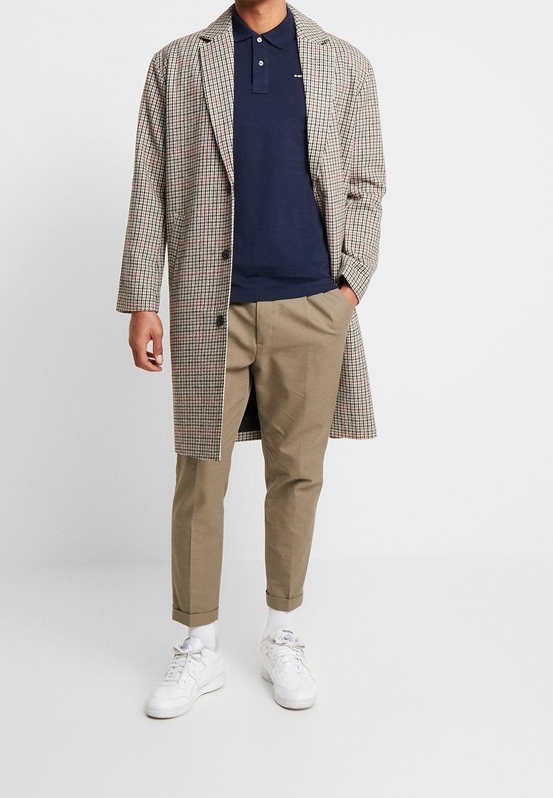 Man wearing navy polo, beige checked long coat, khaki pants, white socks, and white sneakers standing with one hand in pocket.