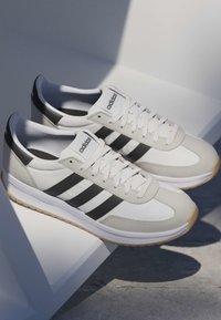 Para białych trampek Adidas z czarnymi paskami i gumowymi podeszwami spoczywających na białej geometrycznej powierzchni w naturalnym świetle.