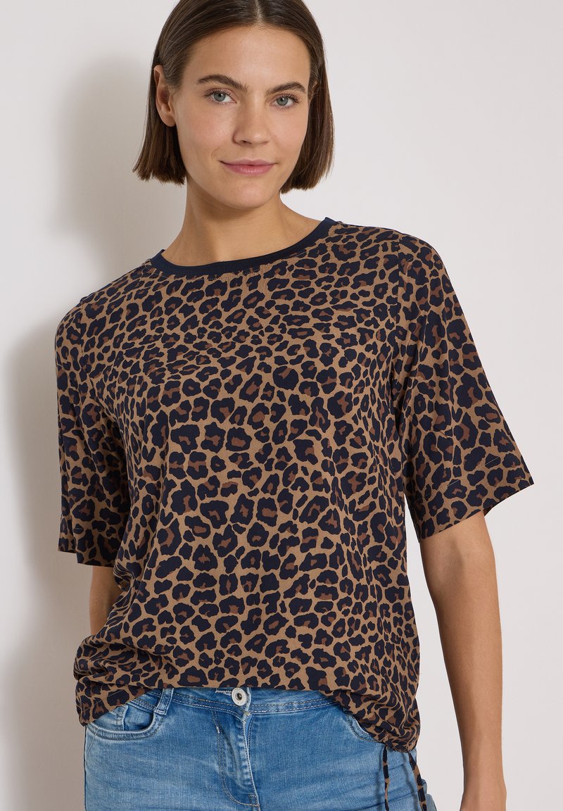 Femme portant une chemise courte à manches courtes avec imprimé léopard marron et noir, rentrée dans un jean bleu, debout devant un fond clair uni.