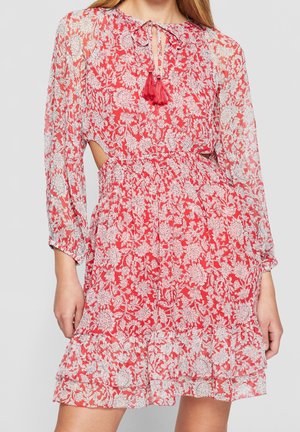 Rode bloemenjurk met lange mouwen en witte bloemmotieven, keyhole-halslijn met rode kwastjes, getailleerde taille en gerufflede zoom, gedragen door een staand persoon.