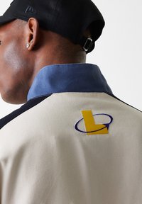 Beige jacket with blue collar and black accents; features an embroidered yellow and purple logo on the back. Textured fabric.