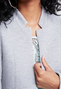 Graue, strukturierte Jacke mit geripptem Muster, mit Stehkragen und offenem Vorderteil, über einem hellblauen Spitzentop getragen.