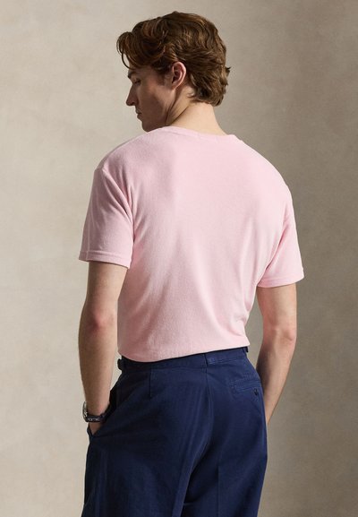 Άνδρας με ανοιχτό καστανά μαλλιά, φορώντας ροζ κοντομάνικο πουκάμισο και σκούρο μπλε παντελόνι, στέκεται με τα χέρια στις τσέπες, βλέποντας προς τα πίσω.