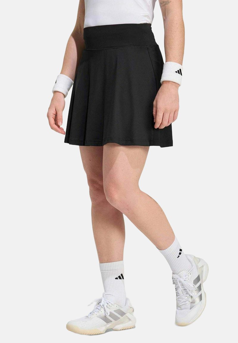 Personne portant une jupe de sport plissée noire, des chaussettes blanches à la cheville, des baskets blanches Adidas et des bracelets blancs avec le logo Adidas.