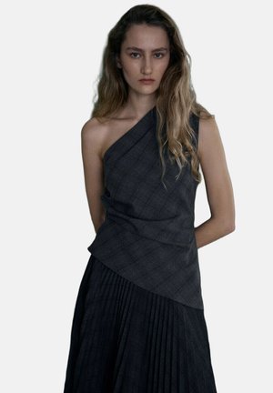 Jonge vrouw met lang golvend haar, gekleed in een donker topje met één schouder en een geplooide rok, staand met handen achter de rug tegen een effen achtergrond.