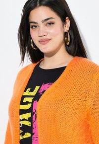 Cardigan orange vif en tricot avec un devant ouvert, porté sur une chemise noire arborant des textes graphiques en jaune et rose.