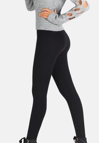 Kobieta ubrana w czarne legginsy, szary sweter z długim rękawem z wycięciami na rękawach oraz czarne botki, stojąca z rękami na biodrach.